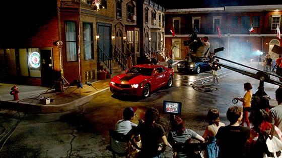满帆大学's Backlot in the evening filled with a full film production crew shooting a scene featuring two muscle cars, 一个红的，一个黑的.